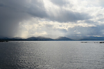 Dramatic clouds cast shadows over tranquil waters near distant mountains during a cloudy afternoon