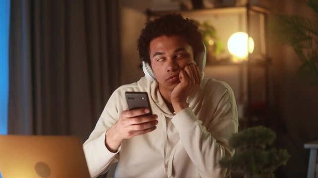 Bored young African American man in headphones staring at smartphone screen at home in the evening after study or work feeling tired distracted or uninterested while browsing or listening to music
