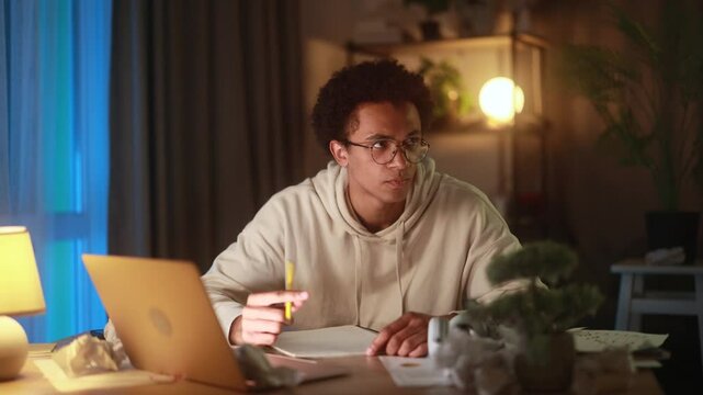 Disappointed young african american man at desk and crossing out ideas in notebook struggling with creative block or frustration while working at home in the evening facing doubt and self-criticism