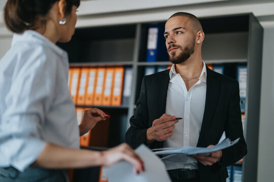 Two business professionals conversing in an office setting, reviewing documents and exchanging ideas. They represent teamwork, collaboration, and professional office culture.