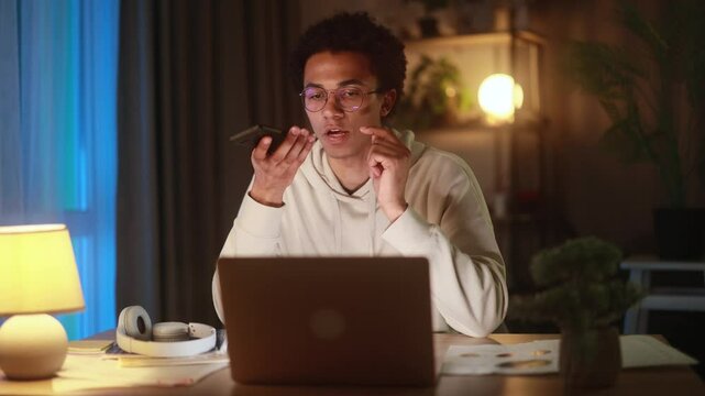 Young African American man in glasses using voice assistant on smartphone at home, talking with serious expression during evening, interacting with AI technology in cozy workspace