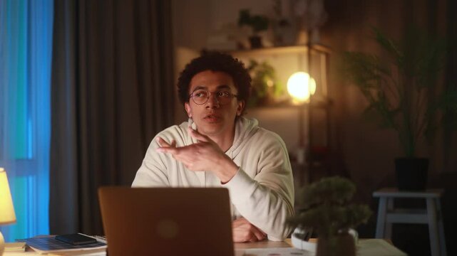 Young man sitting at laptop at home, suddenly getting new idea or insight, raising hand in realisation while working or thinking in cozy evening atmosphere, inspired by creative breakthrough