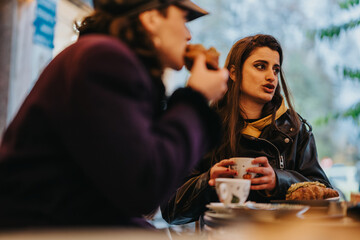 Two females sit at a cafe table, talking and enjoying hot beverages and pastries in an outdoor setting.
