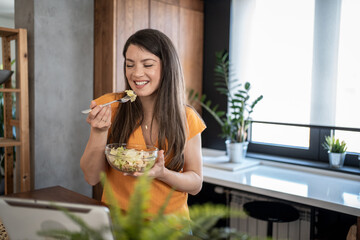 Happy woman eating fresh salad and having video call in kitchen