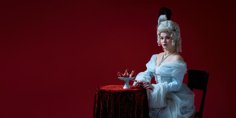 Baroque snack. Royal young woman sitting with bowl of pomegranates, posing calmly against deep red...