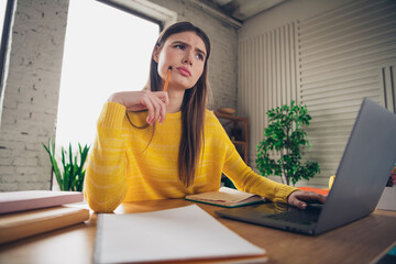 Young woman in a casual yellow sweater thoughtfully working with a laptop indoors in bright natural...