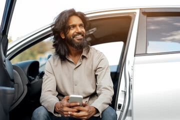 Indian man sitting on the car doorstep with a phone in his hands, happy eastern male smiling and...
