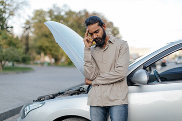 Stressed Indian man standing next to car with open hood, upset young eastern male keeping hand on...
