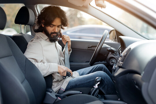 Indian man putting on seatbelt inside the car, focused and careful before driving. Young eastern male sitting in modern vehicle, getting ready before journey, enjoying safe drive