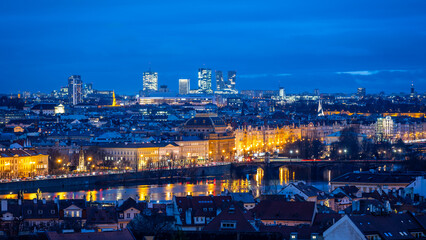 Historic National Theatre stands illuminated beside modern Pankrac buildings. The Prague skyline sparkles at dusk, reflecting along the Vltava River's surface.