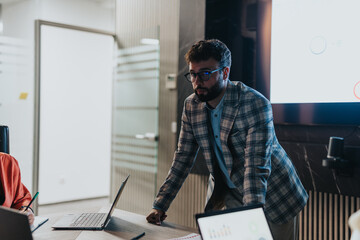 A business professional leans over a conference table, analyzing data from a laptop during a late...