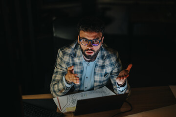 A focused businessman works late, illuminated by a laptop screen in a dark office. The image...