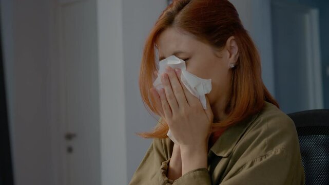 Woman with a migraine sneezes into a napkin while trying to focus on her work in a home office setting. She fidgets, clearly feeling unwell.