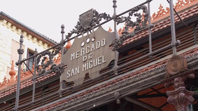 Ornate sign for the mercado de san miguel hanging over the tiled roof of the historic market building in madrid, spain, under a dramatic cloudy sky