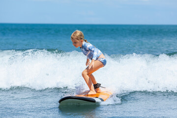 Happy baby girl - young surfer ride on surfboard with fun on sea waves. Active family lifestyle, kids outdoor water sport lessons and swimming activity in surf camp. Summer vacation with child.