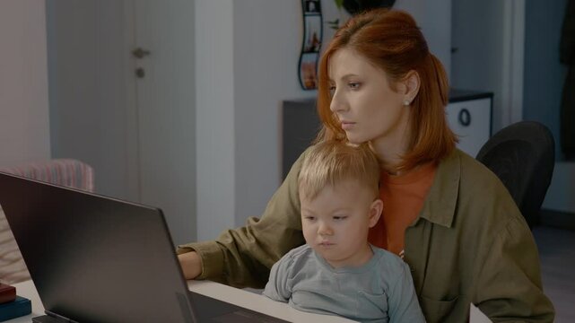 Young mother juggles work on her laptop while holding her baby. She takes notes and analyzes graphs in a cozy home office environment.