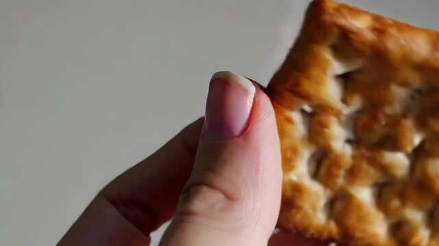 Close-up shot of hands gently breaking apart a textured, golden-yellow cracker or biscuit, revealing its crisp surface under natural light.
