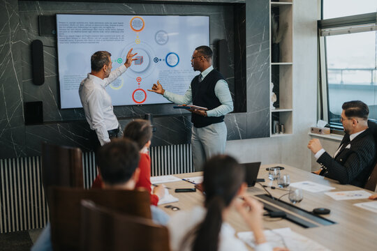 A diverse group of business people engage in a meeting, discussing strategies and data displayed on a large screen in a modern office setting.