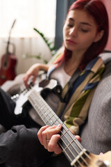 Teenage girl with red hair playing electric bass guitar while sitting in a cozy living room,...