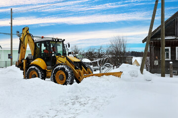 Yellow backhoe loader removes snow near wooden house in rural area.