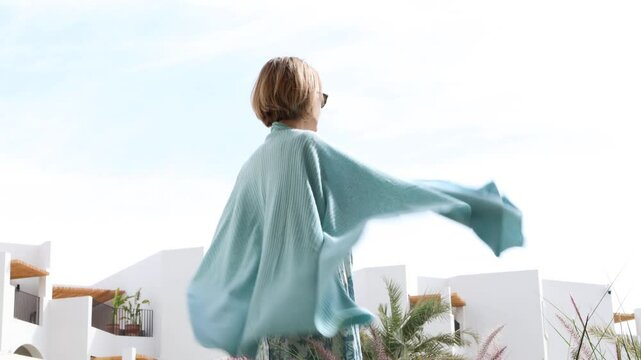 Rear view of woman putting on light blue shawl against sky, warming up on chilly summer day, cozy stylish clothing