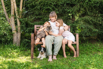 Obraz premium Grandmother sitting on a wooden bench hugging granddaughters with pet cat and dog in a summer garden