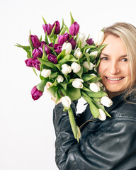 A cheerful blonde woman in a black leather jacket holds a large bouquet of white and purple tulips....