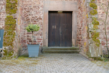 Stone building entrance featuring a wooden door framed by a brick wall. Lush green potted plant enhances the rustic charm, creating a welcoming and serene outdoor scene bad Hersfeld, Germany