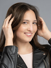Young woman in a model test, close-up shot. Natural expression, soft smile, and lively emotions. Long hair, leather jacket, neutral background. Visible skin texture, soft studio lighting.