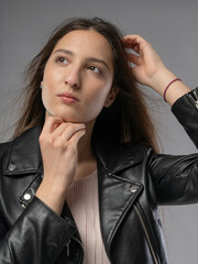 Young woman in a model test, close-up shot. Natural expression, soft smile, and lively emotions. Long hair, leather jacket, neutral background. Visible skin texture, soft studio lighting