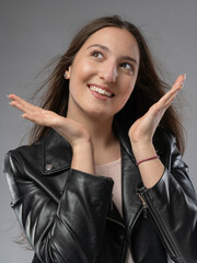 Young woman in a model test, close-up shot. Natural expression, soft smile, and lively emotions. Long hair, leather jacket, neutral background. Visible skin texture, soft studio lighting