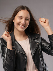 Young woman in a model test, close-up shot. Natural expression, soft smile, and lively emotions. Long hair, leather jacket, neutral background. Visible skin texture, soft studio lighting