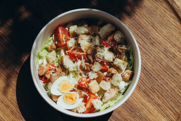 Caesar-style salad with cherry tomatoes, egg halves, croutons and grated cheese in eco-friendly bowl on wooden surface