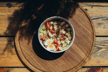 Caesar-style salad with cherry tomatoes, egg halves, croutons and grated cheese in eco-friendly bowl on wooden surface