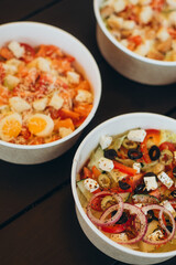 Assortment of colorful fresh vegetable salads in paper bowls on dark wooden table, top view