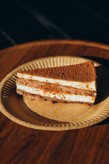 Classic slice of tiramisu cake sprinkled with cocoa powder, served on textured eco paper plate and wooden tray, warm ambient light in rustic cafe setting