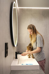 Teen girl preparing to bathe her reluctant cat at the sink in a modern bathroom with soft lighting...