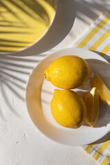 Plate with lemon fruits on the white plaster background with yellow striped pattern napkin with tropical palm leaves shadows. Summer table setting.