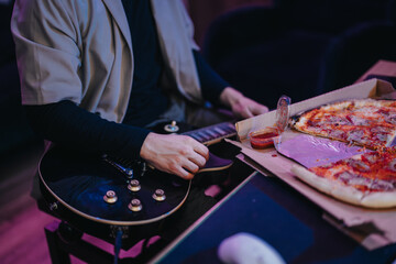 A person unwinds by playing an electric guitar and eating pizza, creating a relaxed atmosphere. The scene combines music with comfort food, highlighting a cozy and enjoyable home setting.