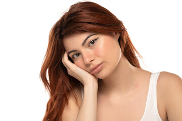 Fototapeta premium Young woman with long red hair looking bored or sad, resting chin on hand. White tank top, white background