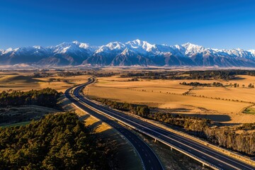 A breathtaking aerial view of a winding road through golden fields, framed by majestic snow-capped mountains under a clear blue sky.