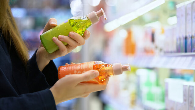 A female customer compares two different shampoo bottles for hair care, reading labels and examining ingredients. Self-care and conscious choice. Close-up of a woman's hands in a cosmetics department 