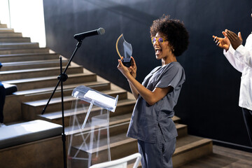 woman in scrubs joyfully receiving award on stage at conference, at conference hall