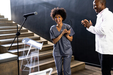 Receiving applause on stage, woman in scrubs smiling, holding glasses, at conference hall