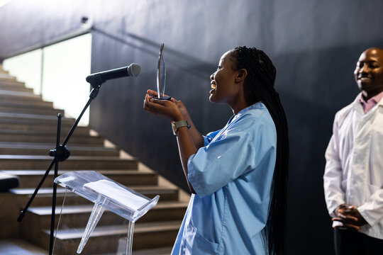 Woman in blue shirt speaking at podium, holding award, smiling confidently, at conference hall