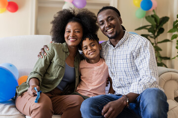 Happy family of three sitting on sofa, celebrating with colorful balloons at home