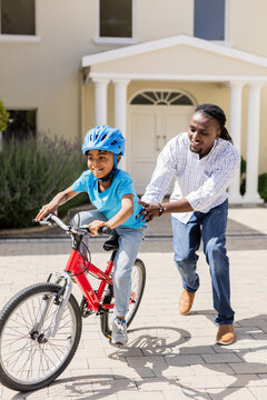 Father teaching son to ride bicycle outside home, both smiling and enjoying