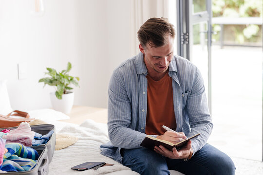 Man writing in notebook while packing suitcase at home, preparing for trip