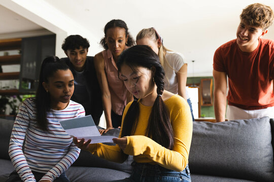 Teen reading college acceptance letter on couch, diverse friends, sharing excitement, at home - Powered by Adobe