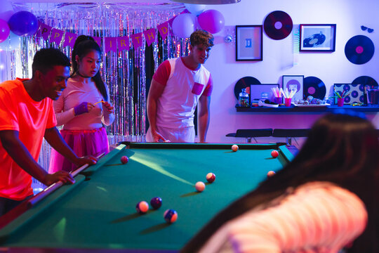 Diverse teenagers playing pool at birthday party, enjoying festive decorations and music, at home